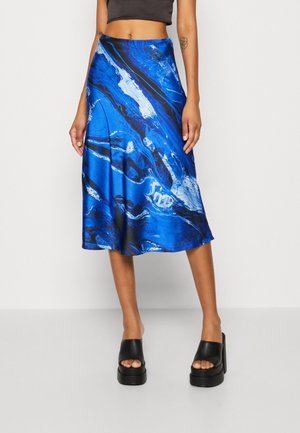 Nainen, joka pukeutunut siniseen ja mustaan abstraktiin printtimidihelmaan, mustaan lyhyeen toppiin ja mustiin korokepohjaisiin sandaaleihin, seisoo yksivärisen taustan edessä.