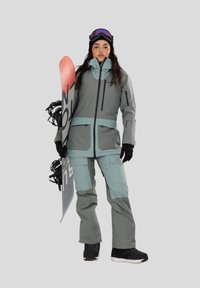 Snowboarder in een grijs en teal geïsoleerd jack en broek, met zwarte handschoenen en laarzen, die een snowboard vasthoudt met een rood en wit ontwerp.