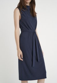 Granatowa sukienka bez rękawów z wysokim kołnierzem i związanym paskiem w talii. Wykonana z gładkiego materiału, o długości do kolan.