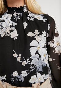 Svart blus med blommönster i vitt och ljusgrått, med smockad halsringning och delikata blommor. Mjuk, genomskinlig tyg.