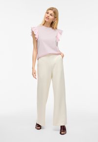 Helles rosa Kurzarmoberteil mit Rüschendetails, kombiniert mit cremefarbenen weiten Hosen und braunen offenen Sandalen. Schlichtes, lässiges Ensemble.
