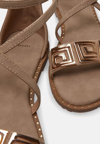 Beige ploché sandály s přeškrtnutými pásky, texturovanou stélkou, zlatými geometrickými akcenty a cvočkovými detaily kolem podešve.