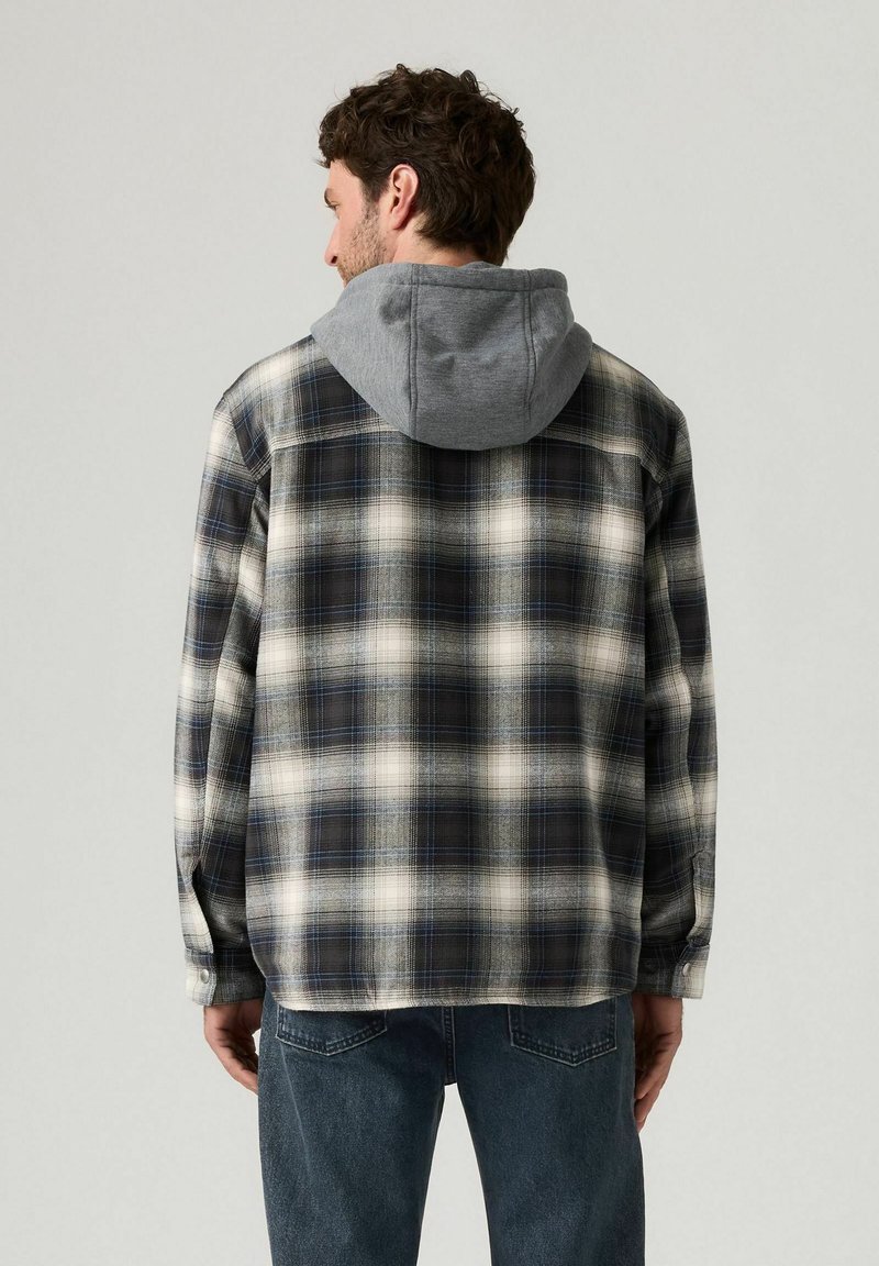 Chemise en flanelle avec capuche grise, présentant un motif écossais en noir, blanc et bleu. Texture douce, manches longues et fermetures à boutons.