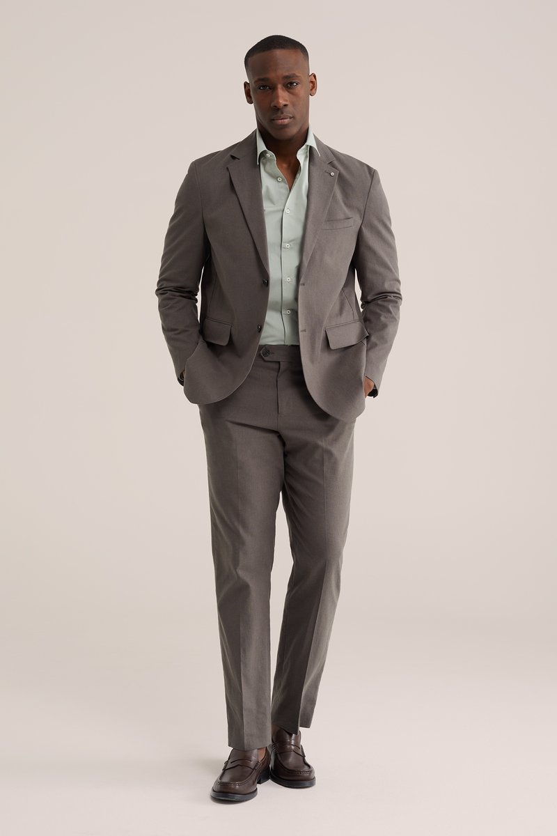 Muž v sivom obleku s mentolovo zelenou košeľou a hnedými mokasínami stojí s rukami v vreckách na jednoliatom pozadí.