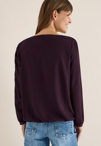 Mørkelilla bluse med lange ærmer og en afslappet pasform, der har en simpel rund halsudskæring og en blød tekstur, parret med lyseblå jeans.