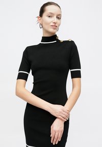 Γυναίκα που φοράει εφαρμοστό μαύρο φόρεμα με λευκές ρίγες στα μανίκια και στον γιακά, χρυσά κουμπιά στον ώμο και ασημένια κρίκους σκουλαρίκια, με λευκό φόντο.