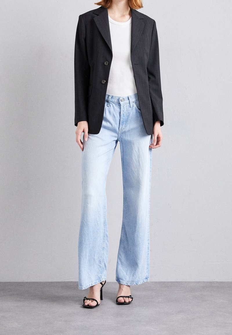 Mustapukuinen räätälöity bleiseri, jossa on kaksi nappia, valkoinen t-paita, vaaleansiniset leveälahkeiset farkut ja mustat korkokengät harmaalla taustalla.