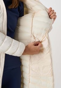 Personne vêtue d'une tenue bleu marine tient ouverte une veste matelassée crème pour montrer sa poche intérieure et sa doublure rembourrée.