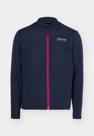 Navy jacket featuring a red zipper, long sleeves, and a stand collar. Includes a logo on the chest and side zippered pockets.