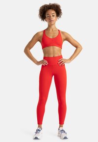 Soutien-gorge de sport rouge et leggings taille haute en tissu extensible. La modèle se tient avec les mains sur les hanches, portant des baskets blanches et bleues.