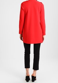 Manteau rouge à manches longues avec une coupe droite, doté d'un petit col et de détails à boutons. Associé à un pantalon noir ajusté et des mules à talons.