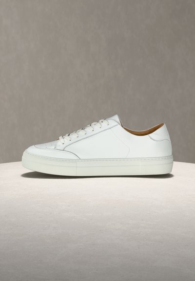 Weißer flacher Leder-Sneaker mit Schnürung vorne, flacher Sohle und perforierten Details an der Zehenpartie, seitlich auf einer neutralen Oberfläche gezeigt.