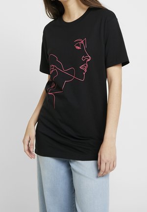 Kobieta w czarnej koszulce z różowym wzorem przedstawiającym ciągłą linię twarzy, w zestawieniu z jasnoniebieskimi dżinsami, na jednolitym szarym tle.