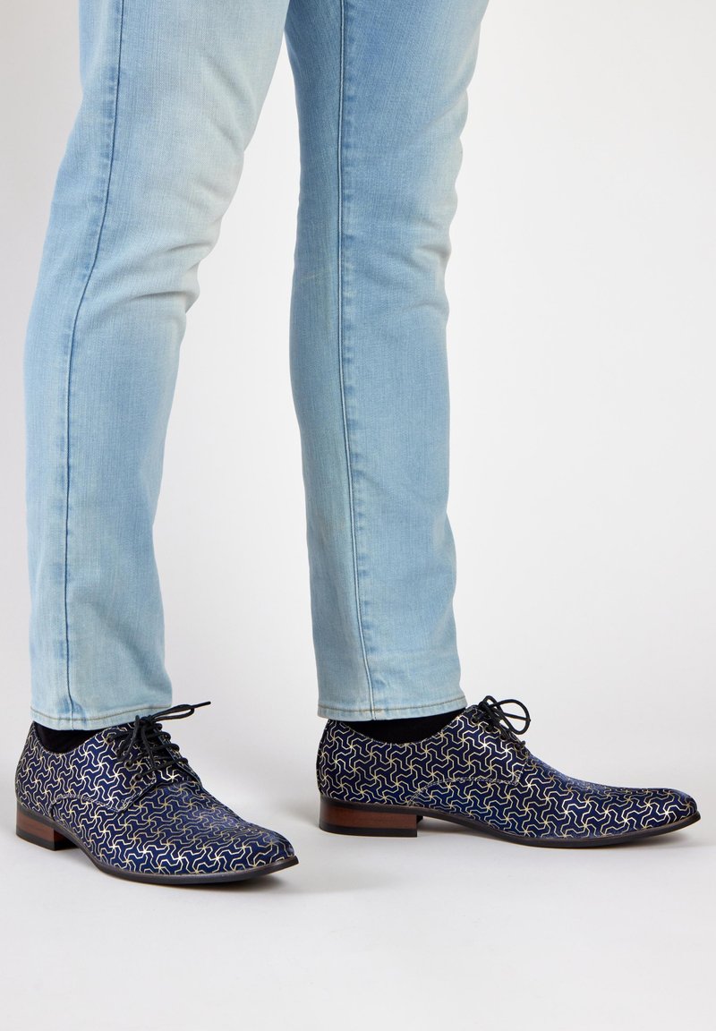 Navy blue shoes with a swirling gold pattern, lace-up design, low heel, and a smooth texture, paired with light blue jeans.
