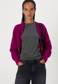 Cardigan magenta a maglia con texture a coste e maniche oversize, indossato sopra una maglietta grigia aderente; abbinato a jeans in denim scuri.