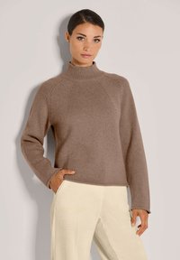 Brauner, gestrickter Pullover mit hohem Kragen, strukturiertem Rippmuster und Raglanärmeln, kombiniert mit hellen Cordhosen.