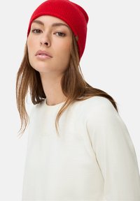 Rode gebreide beanie met een ribbelstructuur en een bruin labeldetail, gecombineerd met een lichtgewicht crèmekleurige sweatshirt met een ronde hals.