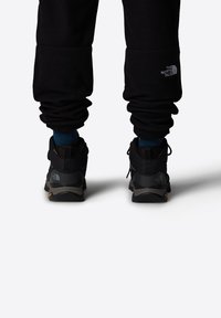 Czarne buty trekkingowe z gumową podeszwą, z szarymi akcentami i logo, połączone z czarnymi spodniami dresowymi i niebieskimi skarpetami.