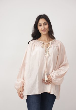 Kobieta o długich ciemnych włosach, ubrana w jasnoróżową bluzkę z koralikami i ciemnoniebieskie dżinsy, stojąca na tle jednolitego, jasnego tła.