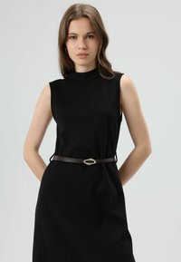 Zwarte mouwloze jurk met een hoge halslijn en geribbelde textuur, getailleerd met een bruine riem met een goudkleurige ovale gesp.