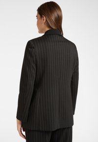 Donna con capelli castani lisci, che indossa un blazer a righe nere e pantaloni abbinati, vista da dietro su uno sfondo semplice.