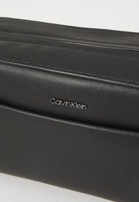 Fekete bőr kézitáska sima textúrával, elegáns dizájnnal és dombornyomott "Calvin Klein" logóval. Diszkrét varrási részleteket mutat be.