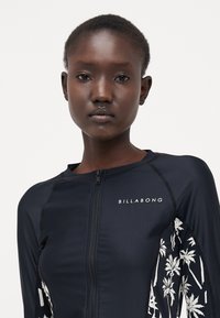 Svart langermet rash guard med glidelås foran. Har hvite palmetre-mønstre på ermene og 'BILLA BONG'-logo på brystet. Glatt tekstur.