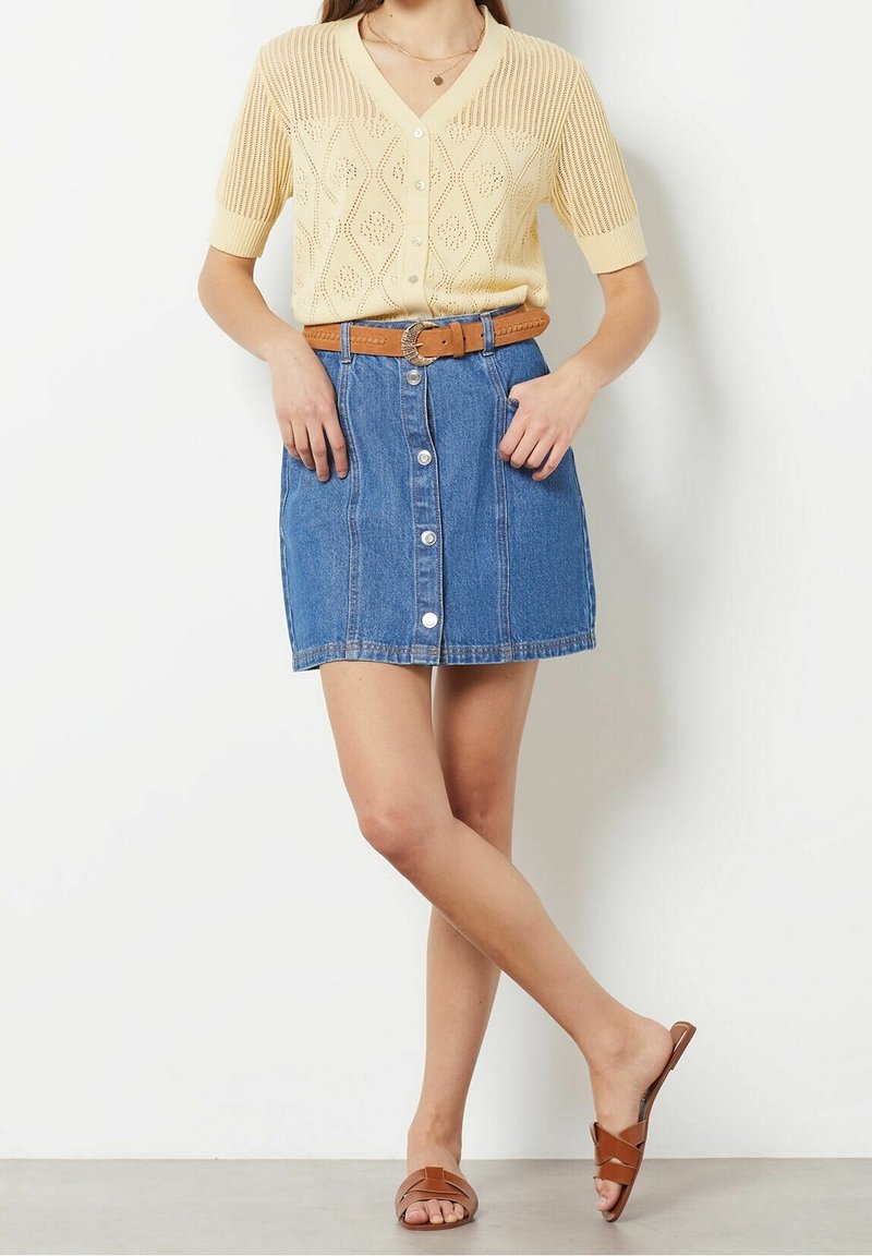 Kremowy sweter na drutach z krótkimi rękawami, zestawiony z niebieską spódnicą dżinsową w kształcie litery A z przodu zapinany na guziki i brązowym paskiem. Brązowe sandały wsuwane.