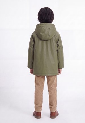 Kind steht mit dem Rücken zur Kamera, trägt eine olivgrüne Kapuzenjacke, beige Hose und braune Schuhe vor einem schlichten weißen Hintergrund.