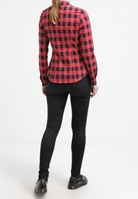 Camicia a quadri rossa e nera con maniche lunghe, abbinata a jeans skinny neri e scarpe nere. Design visibile sul retro e vestibilità su misura.