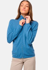 Femme portant un sweat à zip bleu clair et un pantalon beige, fermant le col du sweat, se tenant devant un fond clair uni.