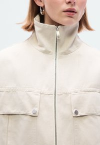 Φωτεινό μπεζ μπουφάν με ψηλό γιακά, μπροστινό φερμουάρ και δύο τσέπες στο στήθος με ασημί διακοσμητικά στοιχεία. Υφή λείου βαμβακερού υφάσματος.