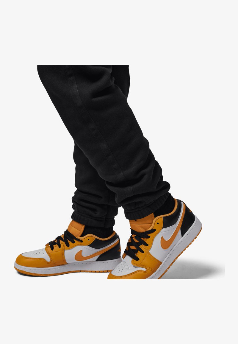 Sneakers Nike w kolorach pomarańczowym, czarnym i białym, wykonane ze skóry, z teksturowaną cholewką, wyraźnym swooshem i czarnymi sznurówkami. Noszone są czarne dresy.