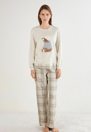 Lichtbeige sweatshirt met een geborduurd berenmotief in een blauwe trui, gecombineerd met plaid pyjamabroek in beige, blauw en bruine tinten.