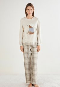 Sudadera beige clara con un oso bordado que lleva un suéter azul, combinada con pantalones de pijama de cuadros en tonos beige, azul y marrón.