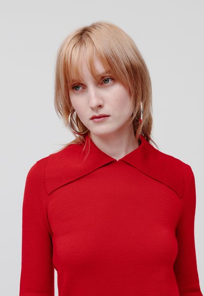 Pulover tricotat roșu cu un guler lat și plat și țesătură texturată. Modelul are părul scurt și blond și cercei mari în formă de inel. Fundal neutru.