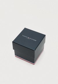 Tmavomodrá štvormilimetrová škatula s vekom, na spodnom okraji s logom "TOMMY HILFIGER" v bielej, červenej a modrej farbe.