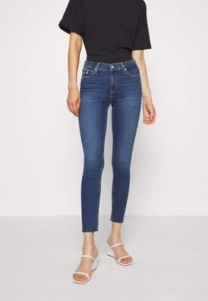 Osoba nosząca ciemnoniebieskie obcisłe dżinsy z surowym wykończeniem, czarny krótki top oraz białe sandały na obcasie, stojąca na jasnym tle.