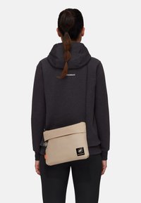 Hellbeige Umhängetasche mit einem umklappbaren Deckel, strukturierten Oberflächen, schwarzem Logo-Patch und verstellbarem Riemen, getragen über einem dunkelgrauen Hoodie.