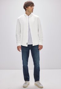 Weißes langärmeliges Button-Up-Hemd über einem weißen T-Shirt, kombiniert mit dunkelblauen Jeans und weißen Turnschuhen. Einfaches Design, Knopfdetails sichtbar.
