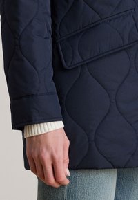 Nærbilde av en hånd ved siden av en marineblå quiltet jakke med buede sømmer, klaffelomme og en hvit ribbestrikket mansjett synlig over lyseblå jeans.