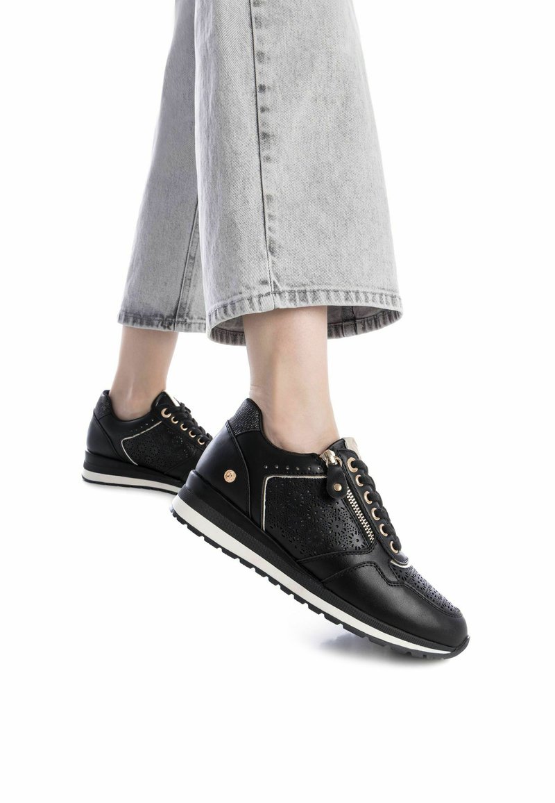 Zapatillas negras con diseño perforado, suela blanca y detalle de cremallera, combinadas con unos vaqueros cropped grises. Presentan acentos dorados y cierre con cordones.