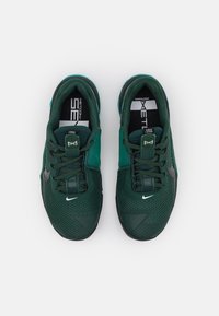 Dvojice tmavozelených tenisek Nike Metcon 7 s síťovinovým svrškem, černým logem swoosh a zajištěnými tkaničkami, zobrazených shora na bílém pozadí.