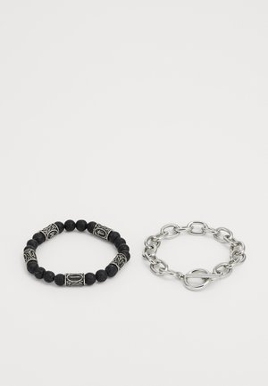 Bracciale di perle nere con elementi decorativi in argento accanto a un bracciale in argento con chiusura a toggle. I materiali testurizzati sono evidenti.