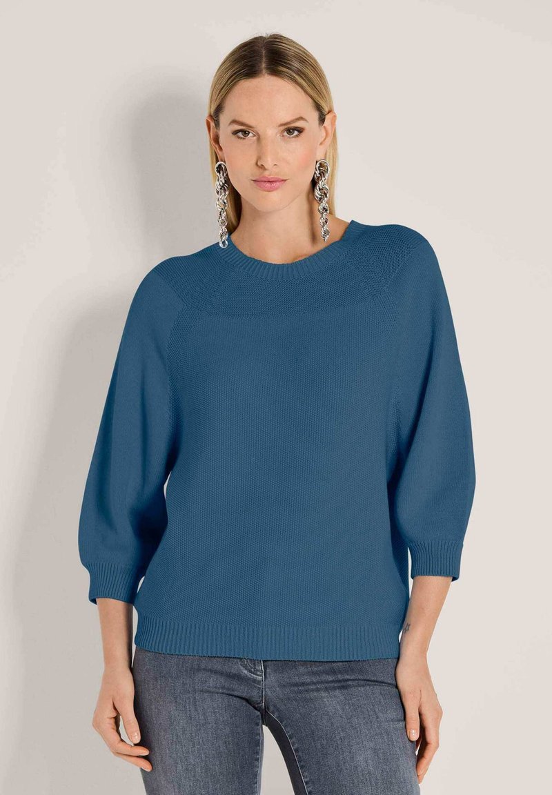 Eine Frau trägt einen blauen Strickpullover mit Dreiviertelärmeln und großen silbernen Kettenohrringen und steht vor einem schlichten Hintergrund.