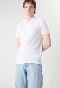 Young man wearing a white short-sleeve polo shirt and light blue jeans, standing against a plain light background with hands behind back.