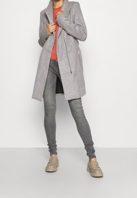Persona che indossa un lungo cappotto grigio su un maglione arancione, jeans attillati grigi e mocassini beige, in piedi di fronte a uno sfondo bianco uniforme.