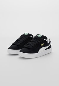 Czarne zamszowe sneakersy z białymi gumowymi podeszwami, z grubymi czarnymi sznurowadłami i białym akcentem w postaci paska PUMA. Złote logo na boku.