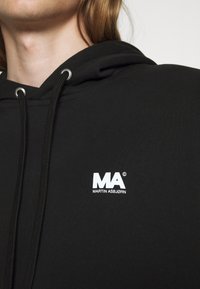 Sort hættetrøje med en glat overflade, med et hvidt logo "MA" og brandnavnet "MARTIN ASBJØRN" broderet på brystet.