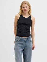 Blond kvinne iført en ermeløs svart topp og løstsittende blå jeans, stående mot en ensfarget hvit bakgrunn.